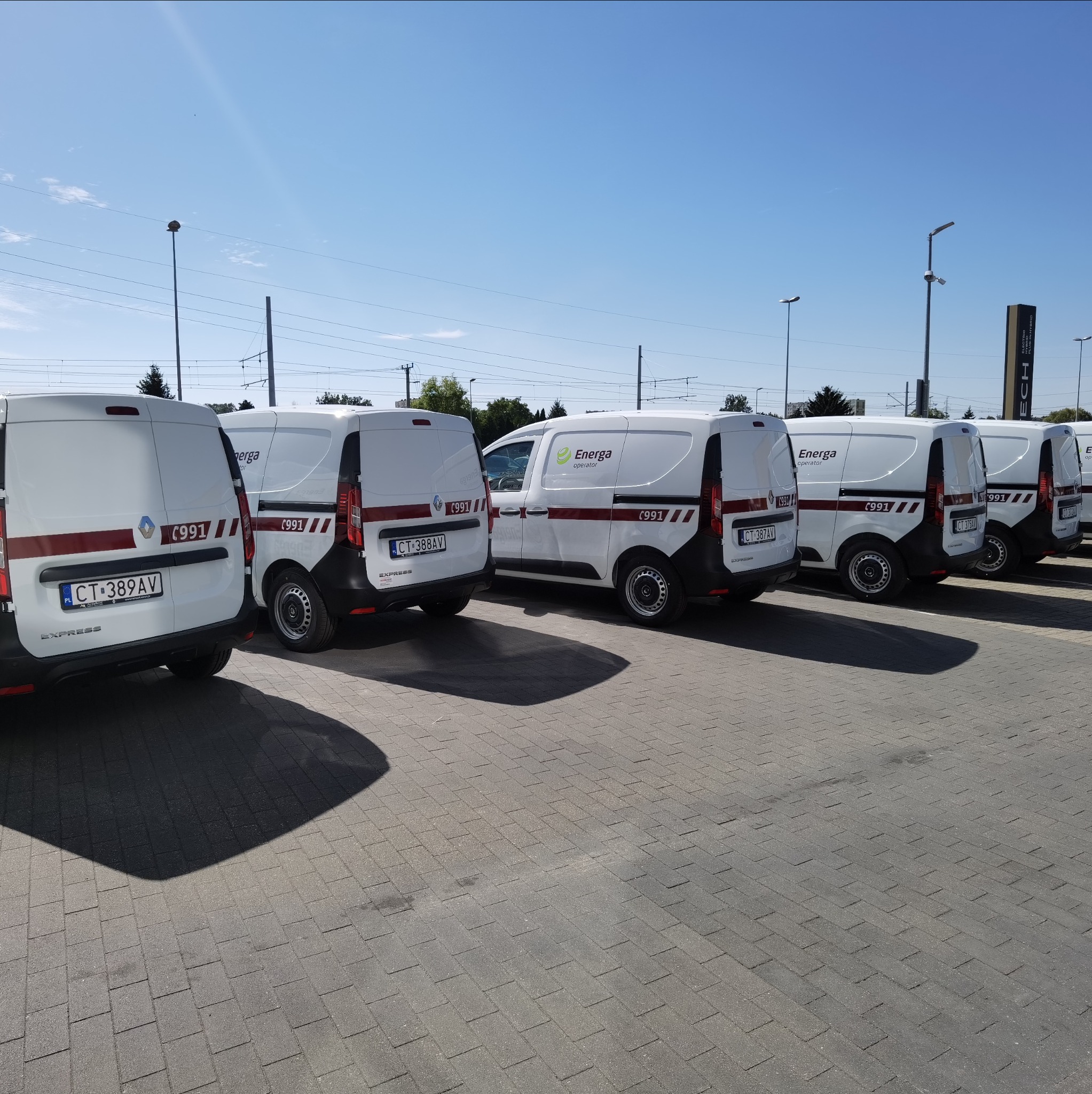 Flota białych vanów z logo Energa i numerem 991, zaparkowanych na placu z kostki. Pojazdy z czerwonym pasem, widoczne tablice rejestracyjne. Słoneczny dzień.