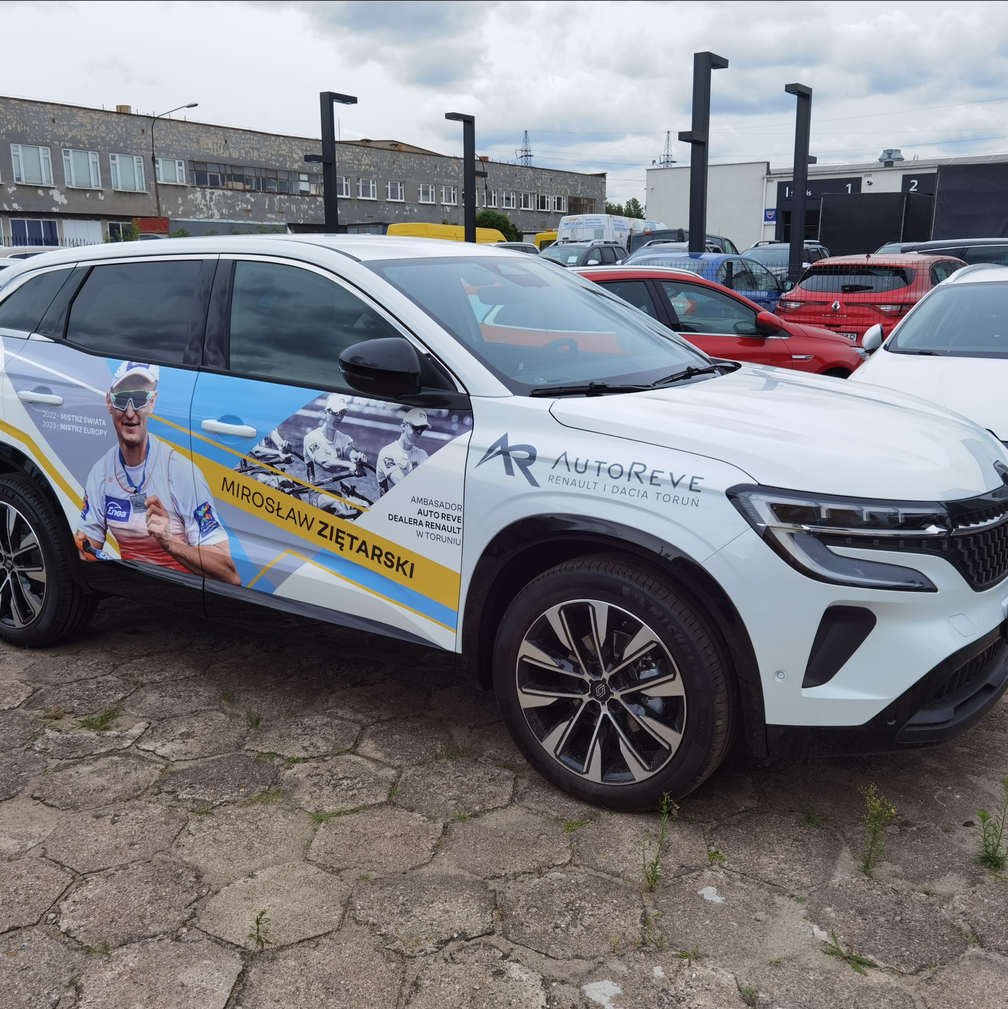 Biały samochód z oklejeniem reklamowym, przedstawiającym sportowca Mirosława Ziętarskiego i logo dealera Renault/Dacia. Reklama sportowca na aucie.