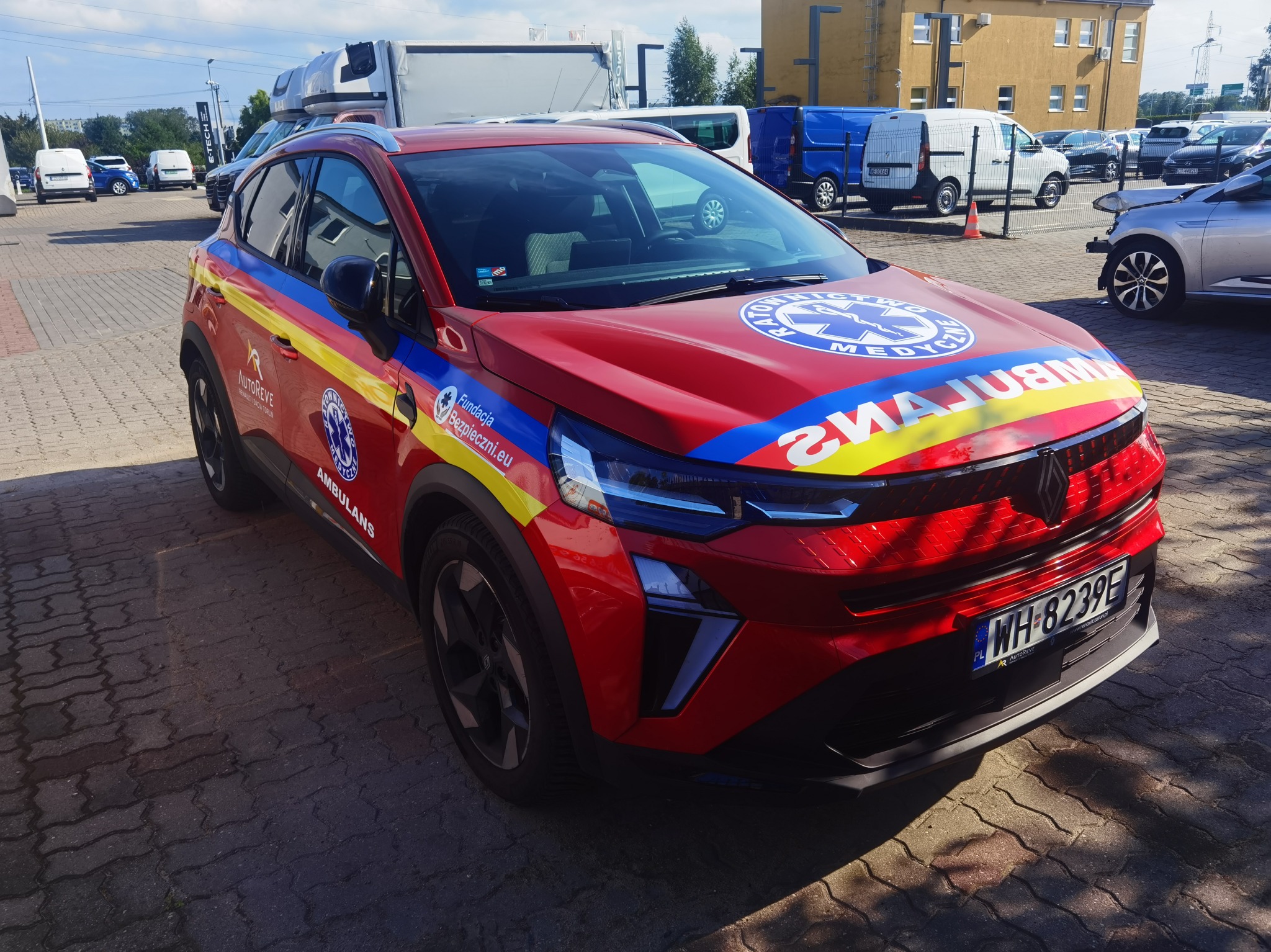 Czerwony samochód z oznaczeniami ambulansu, zaparkowany na brukowanym placu. Widoczne logo i napisy Fundacji Bezpieczni.eu oraz oznaczenia medyczne.