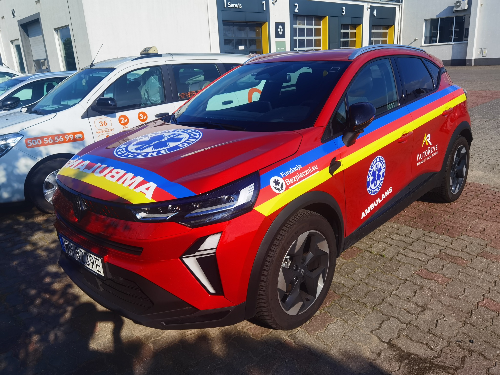 Czerwony samochód medyczny z grafiką ratowniczą i logotypami, zaparkowany na kostce brukowej przed budynkiem serwisu Renault/Dacia, w słoneczny dzień.