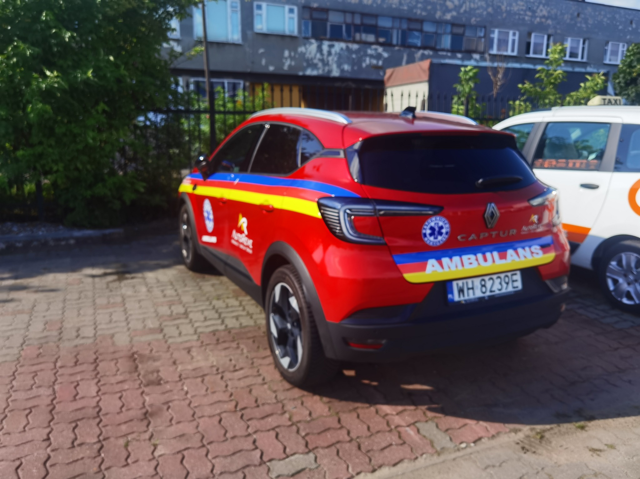 Czerwony Renault Captur z oznaczeniami ambulansu, zaparkowany na brukowanym placu, widoczny bok i tył pojazdu. Obok biała taksówka.