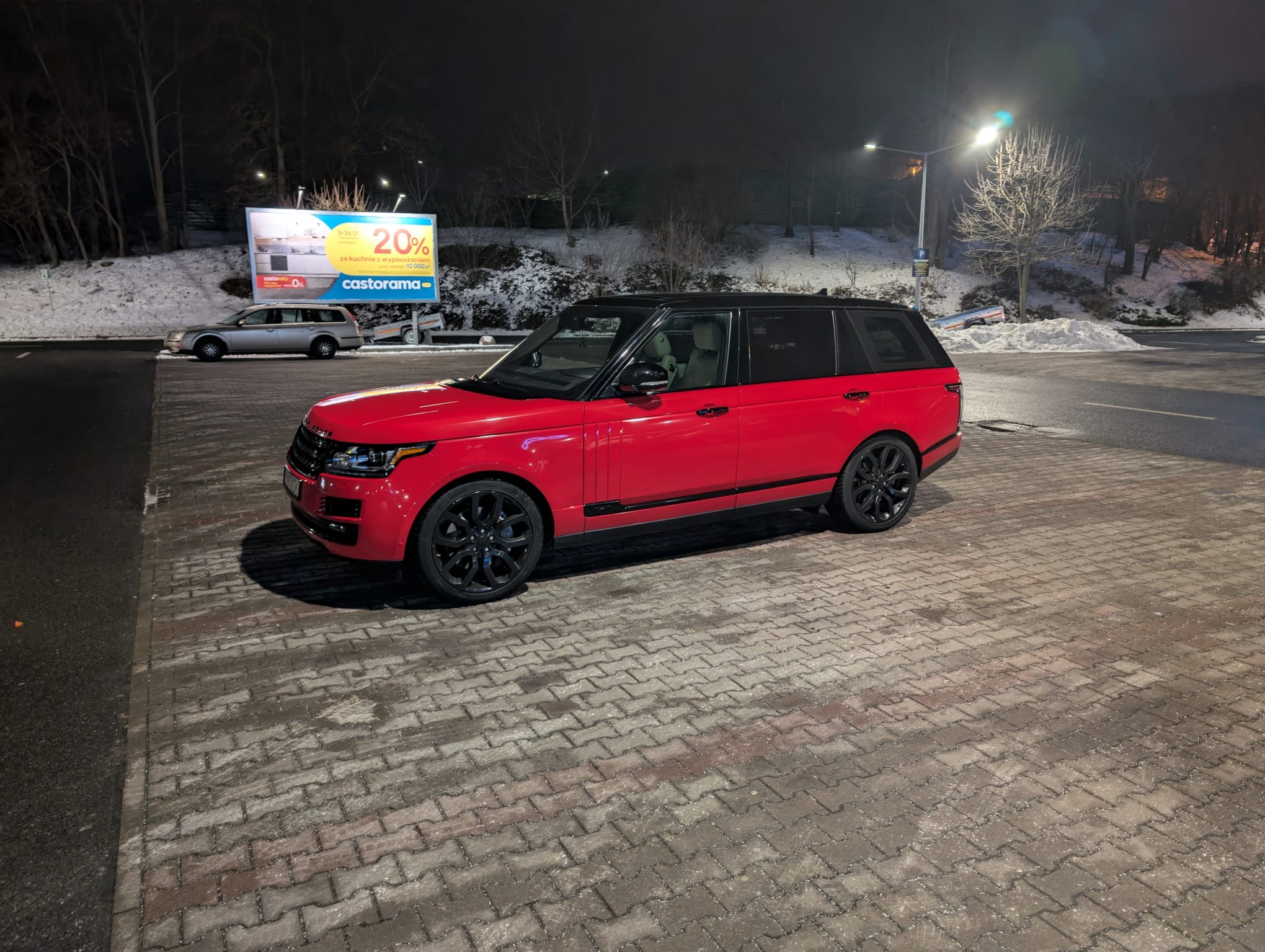 Czerwony Range Rover z czarnym dachem i felgami, zaparkowany nocą na kostce brukowej, w tle billboard Castorama i zasnieżony teren.