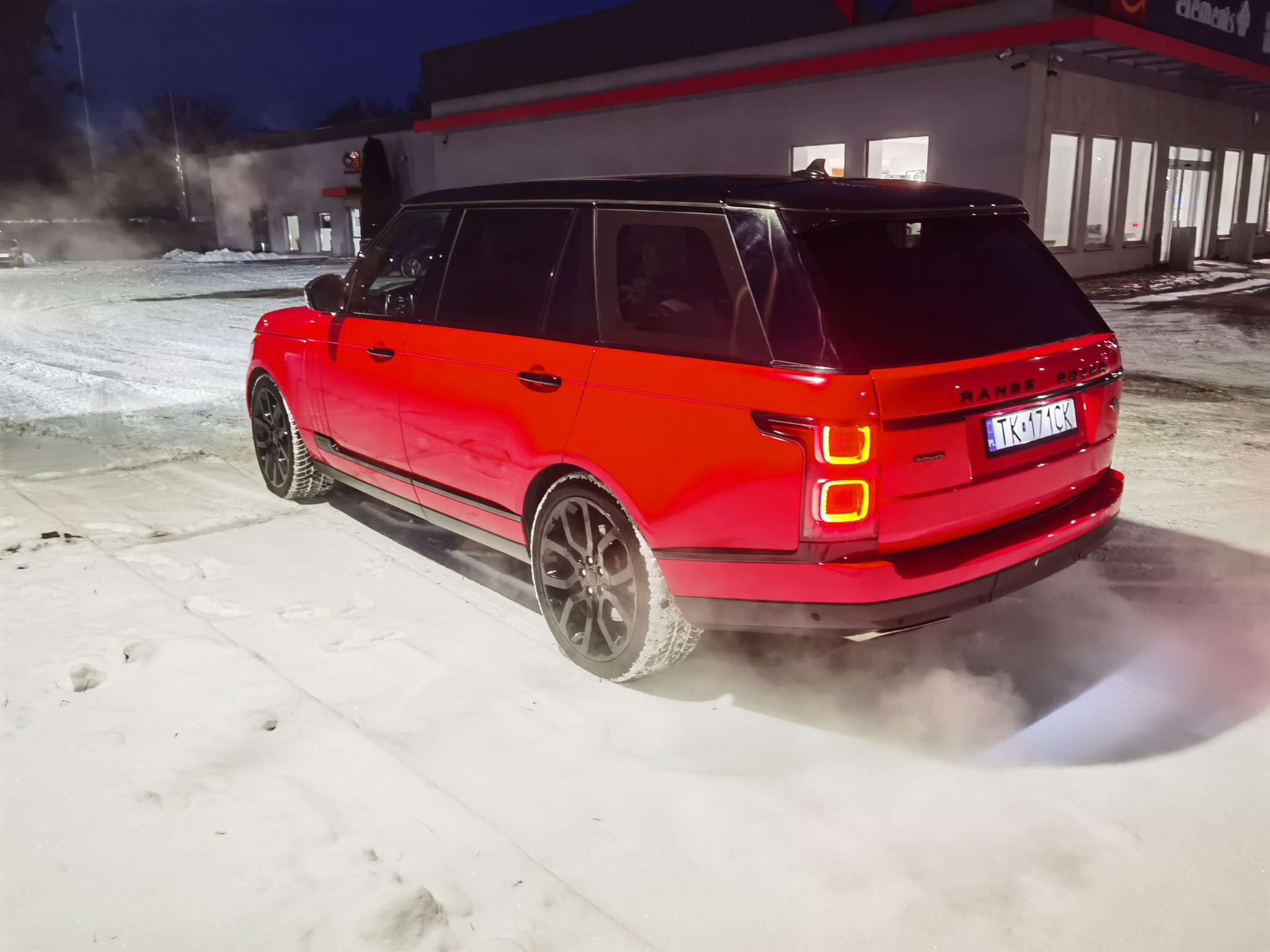 Czerwony Range Rover z czarnym dachem, widziany z tyłu na śniegu, z widoczną tablicą rejestracyjną i światłami. Tło stanowi budynek z oknami.