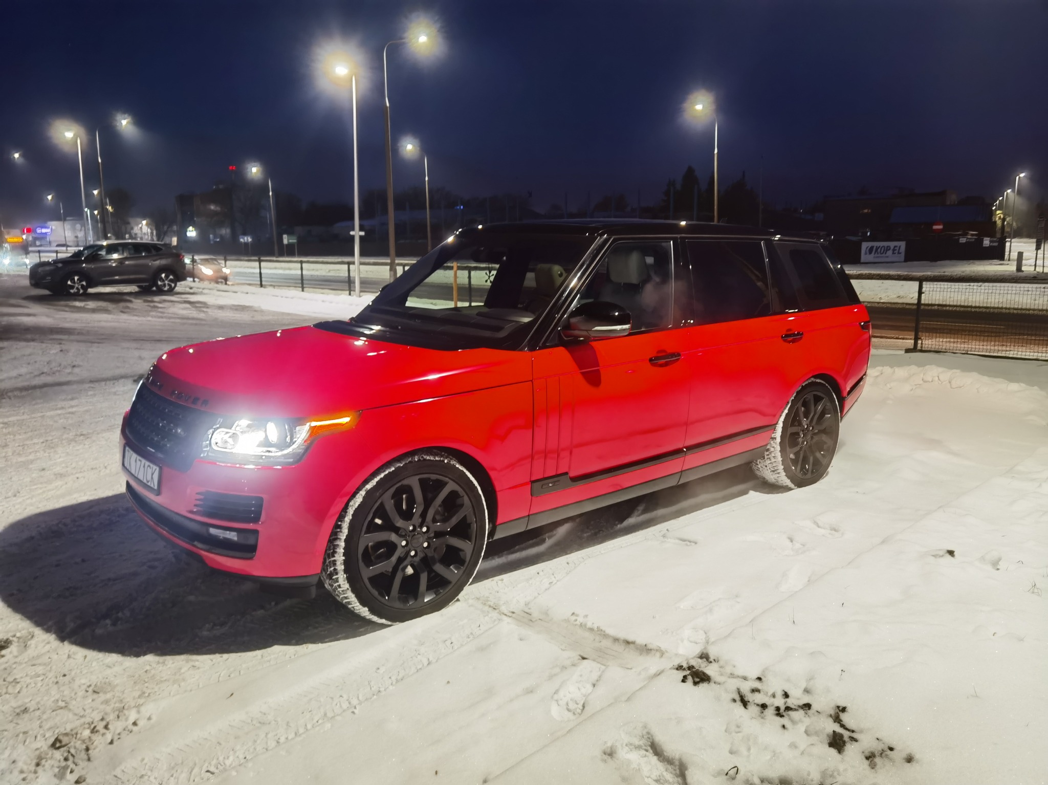 Czerwony Range Rover z czarnymi felgami, zaparkowany na śniegu nocą, oświetlony latarniami ulicznymi. Widoczny numer rejestracyjny.