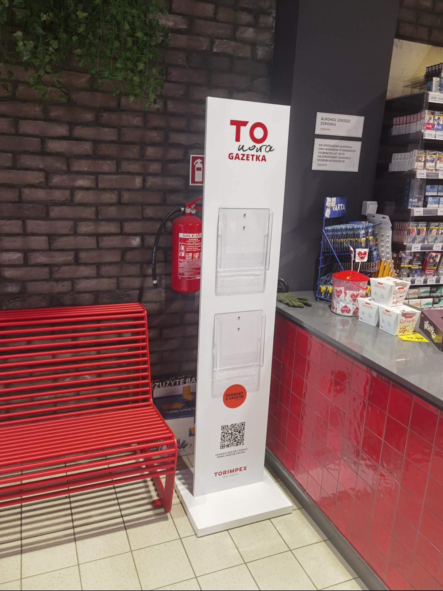 Biały stojak reklamowy z logo 'TO i nowa GAZETKA' i kieszeniami na ulotki, umieszczony w sklepie Torimpex, obok czerwonej ławki i gaśnicy.