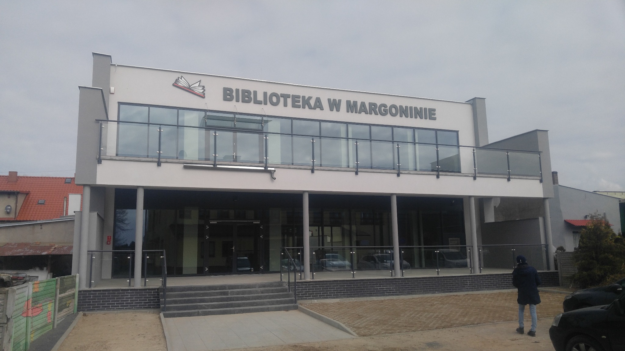 Dwukondygnacyjny budynek Biblioteki w Margoninie z dużymi oknami i szklanymi balustradami, widok z zewnątrz w pochmurny dzień.
