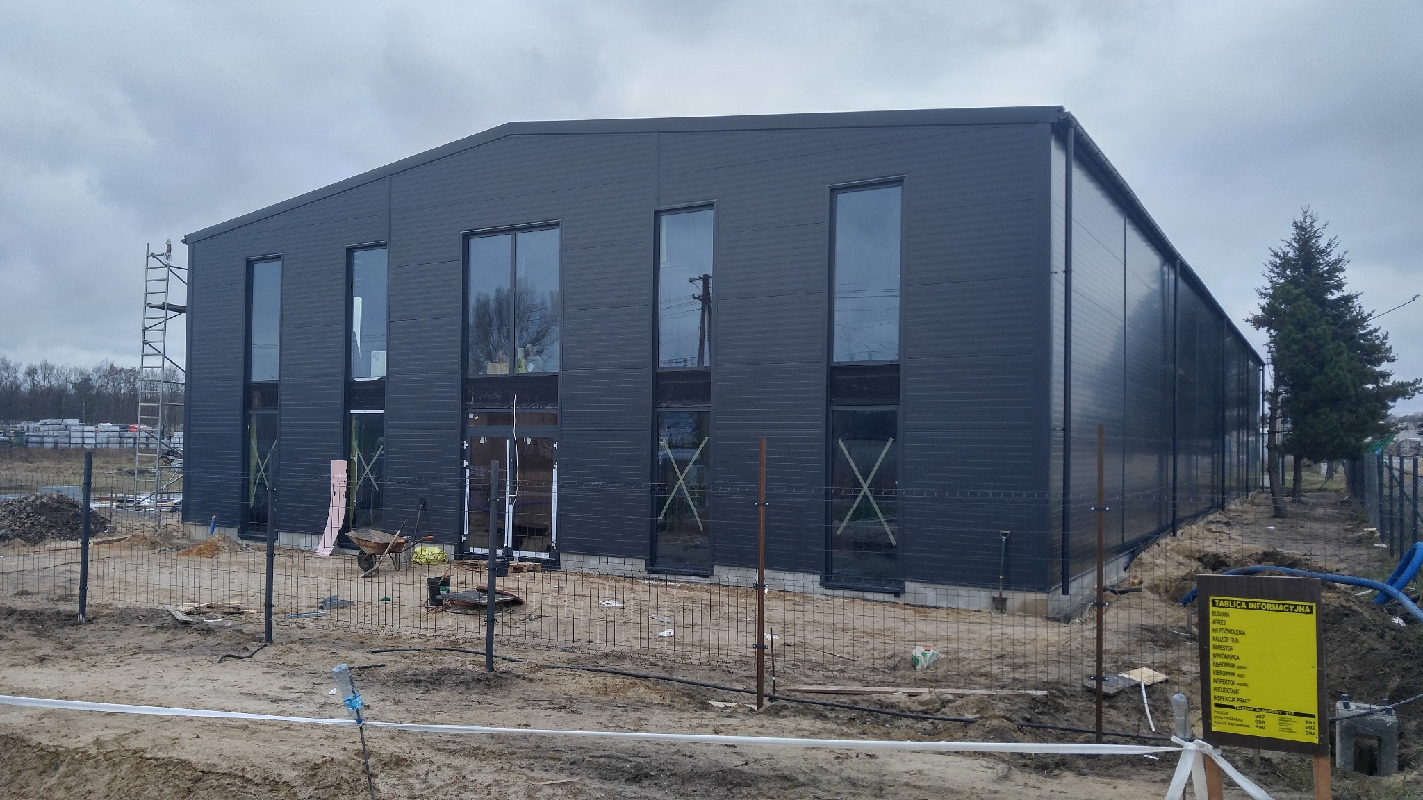 Nowoczesna hala przemysłowa z ciemnoszarej blachy trapezowej, w trakcie budowy, widoczne rusztowania, tablica informacyjna i ogrodzenie placu budowy.