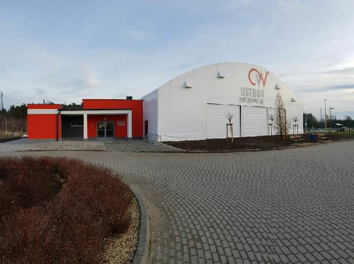 Widok zewnętrzny hali sportowej z białego materiału plandekowego z czerwonym budynkiem administracyjnym połączonym z halą, logo 'OV' na hali, kostka brukowa na pierwszym planie.