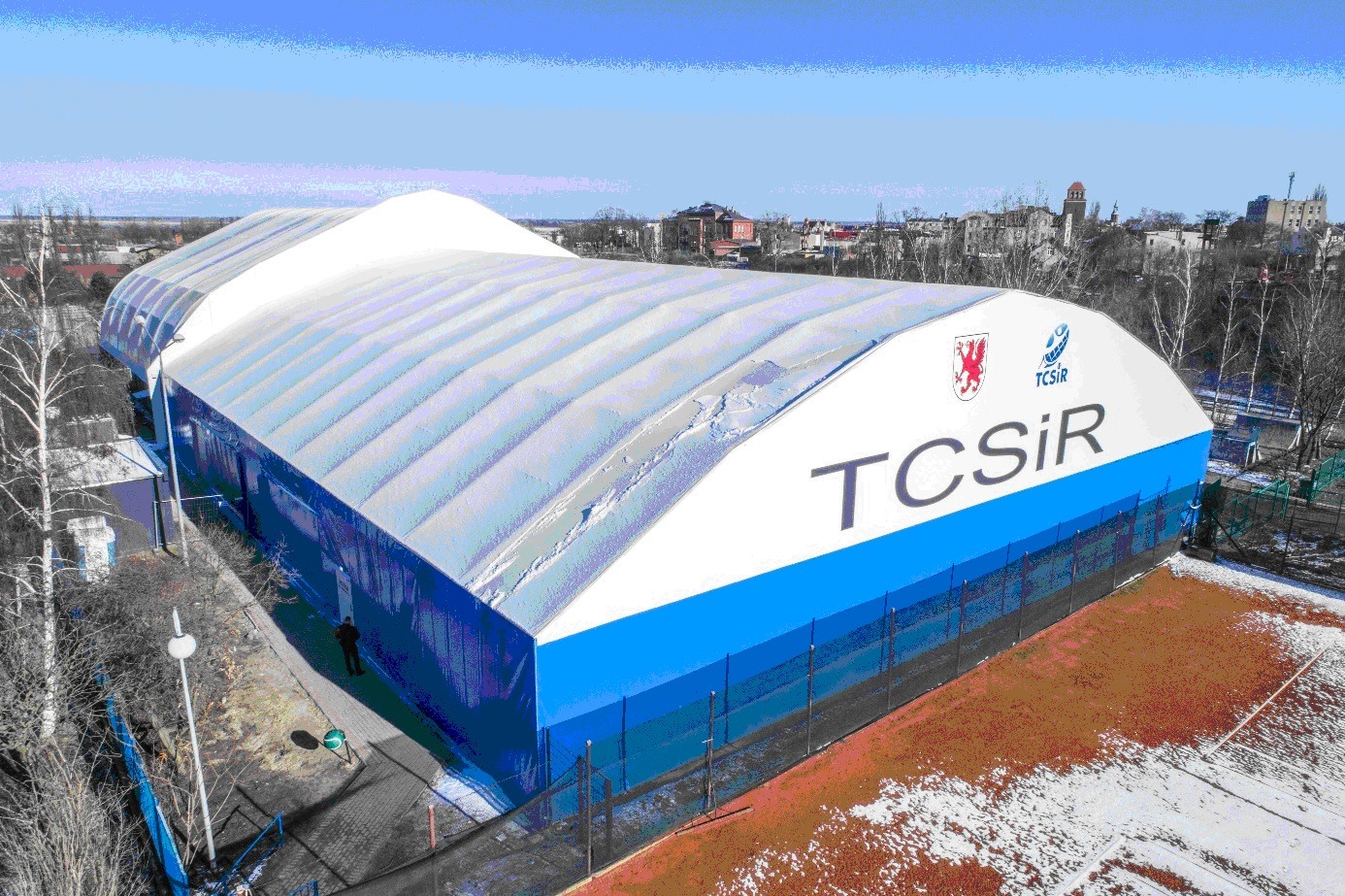 Zadaszony kort tenisowy z białym dachem i niebieskimi ścianami, widok z góry, w tle zabudowa miejska i błękitne niebo.