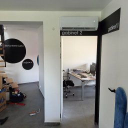 Ciepło-Zimno - Wnętrze biura z zamontowanym klimatyzatorem Gree nad drzwiami do gabinetu, widoczne również schody, kartony i narzędzia na podłodze.