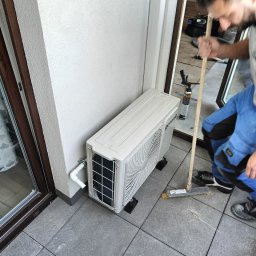 Ciepło-Zimno - Biała jednostka zewnętrzna klimatyzacji Gorenje na balkonie z szarymi płytkami, monter w kombinezonie sprząta resztki po montażu