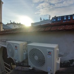 Ciepło-Zimno - Dwa klimatyzatory marki GREE zamontowane na zewnętrznej ścianie budynku z widoczną dachówką i budynkami w tle, oświetlone wschodzącym słońcem.