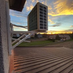 Ciepło-Zimno - Zewnętrzna jednostka klimatyzatora zamontowana na białym wsporniku na tle zachodzącego słońca i widoku na okoliczne budynki.