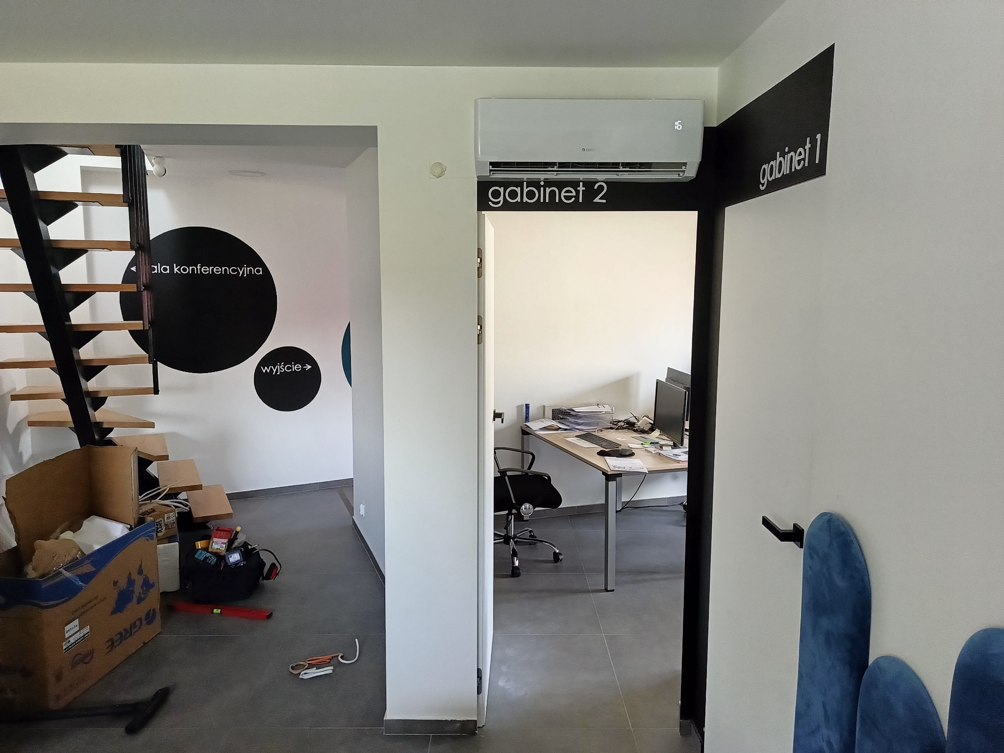 Wnętrze biura z zamontowanym klimatyzatorem Gree nad drzwiami do gabinetu, widoczne również schody, kartony i narzędzia na podłodze.
