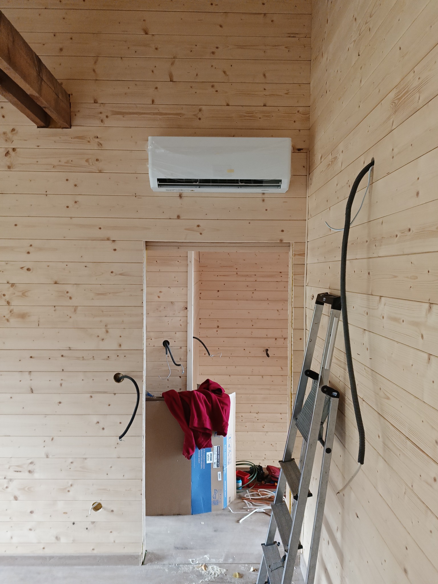 Klimatyzator w folii ochronnej zamontowany na drewnianej ścianie, widoczna drabina i przewody elektryczne, wnętrze w trakcie wykańczania.
