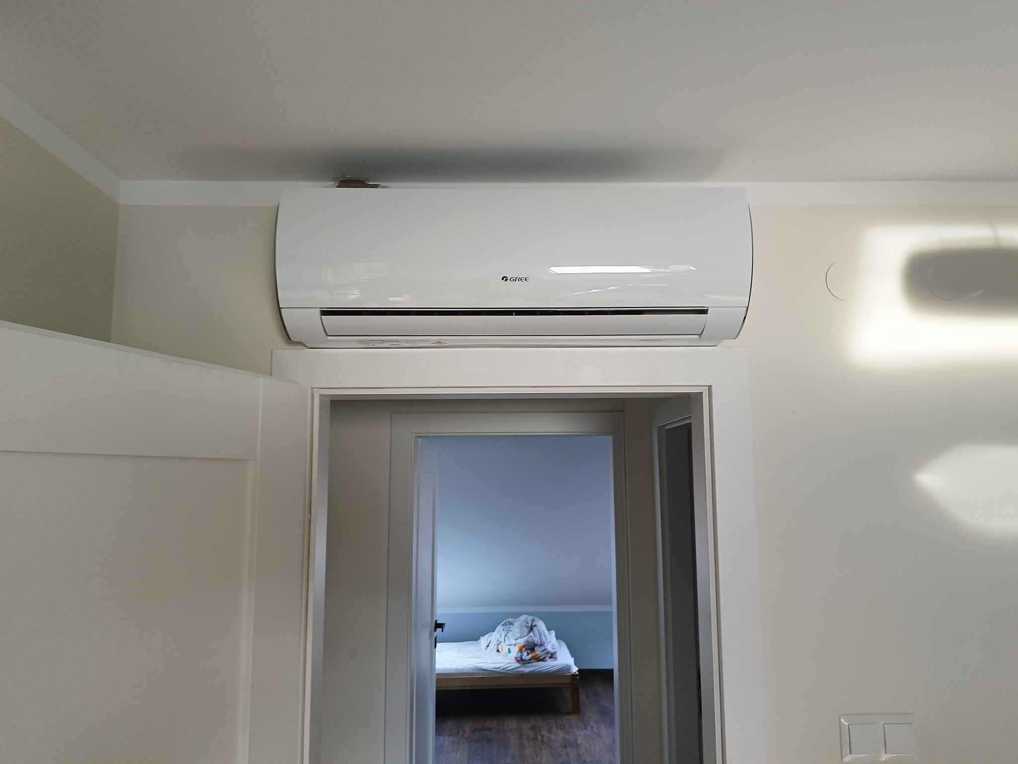 Biały klimatyzator marki Gree zamontowany nad drzwiami do sypialni na poddaszu, widoczne ślady po starym urządzeniu na suficie.
