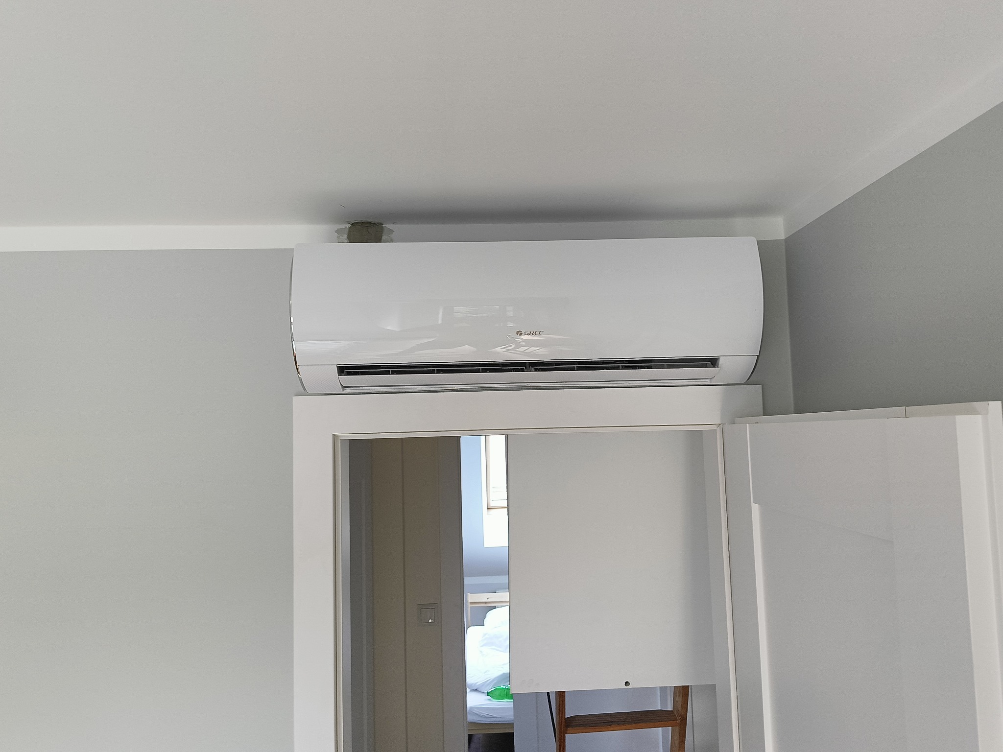 Biała jednostka klimatyzacji marki Gree zamontowana nad drzwiami w pokoju o szarych ścianach i białym suficie, widoczna niedoróbka w postaci szpachli przy suficie.