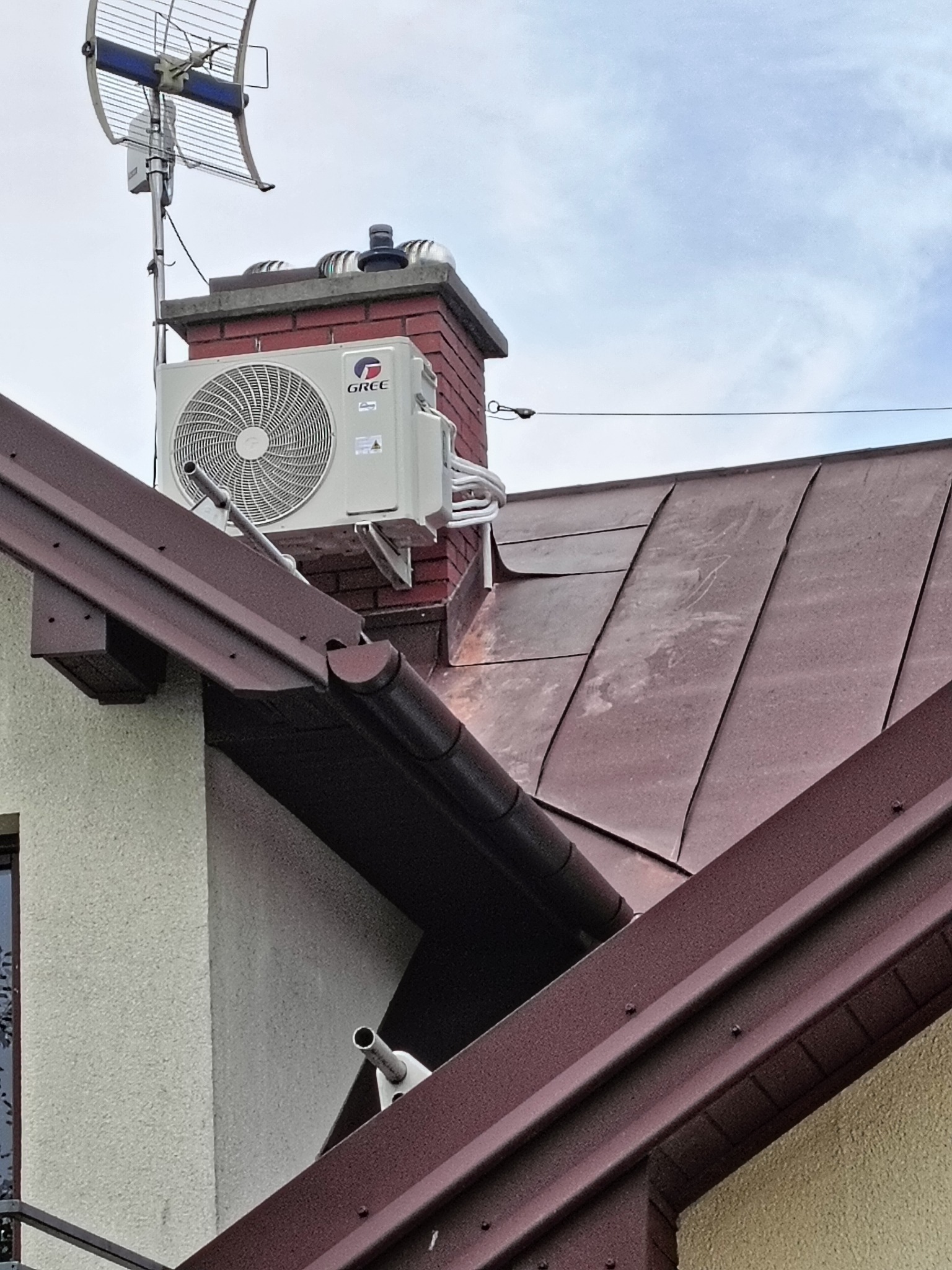 Klimatyzator marki Gree zamontowany na wspornikach przy kominie na dachu domu z brązową blachą, widoczna antena telewizyjna.