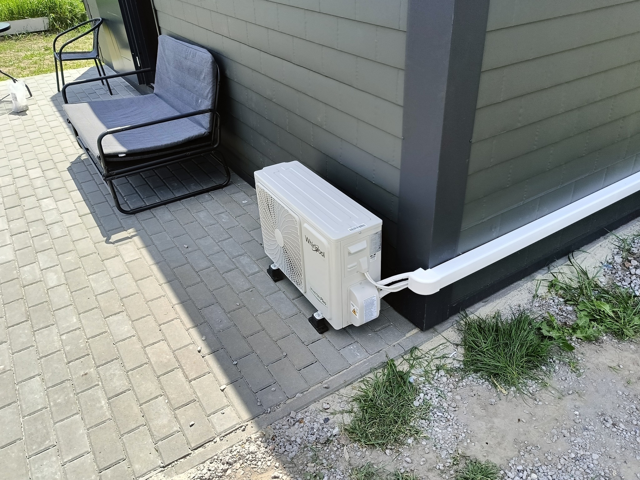 Zewnętrzna jednostka klimatyzacji Whirlpool Inverter Plus zamontowana na kostce brukowej przy szarej elewacji domu, z widoczną białą rurą odprowadzającą skropliny i fragmentem szarej sofy ogrodowej...