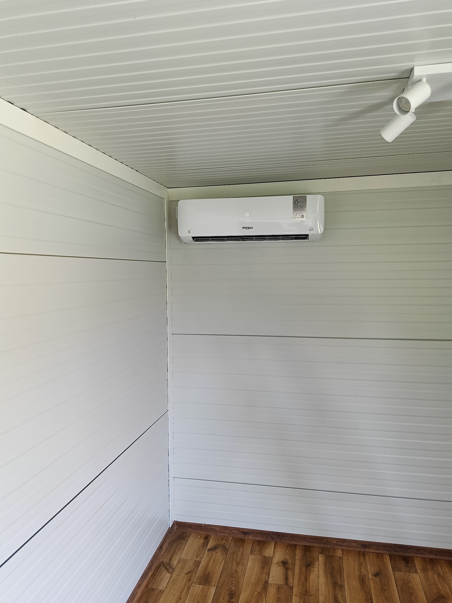 Biały klimatyzator Whirlpool zamontowany na ścianie z paneli w pomieszczeniu z laminowaną podłogą i dwoma reflektorami sufitowymi.
