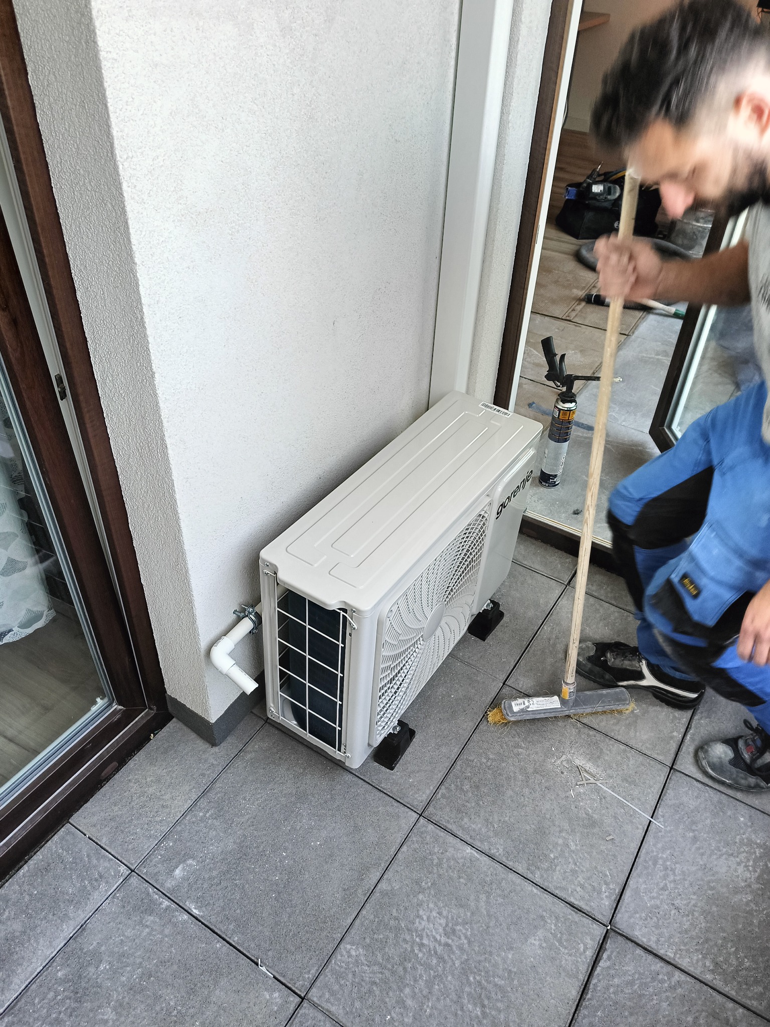 Biała jednostka zewnętrzna klimatyzacji Gorenje na balkonie z szarymi płytkami, monter w kombinezonie sprząta resztki po montażu