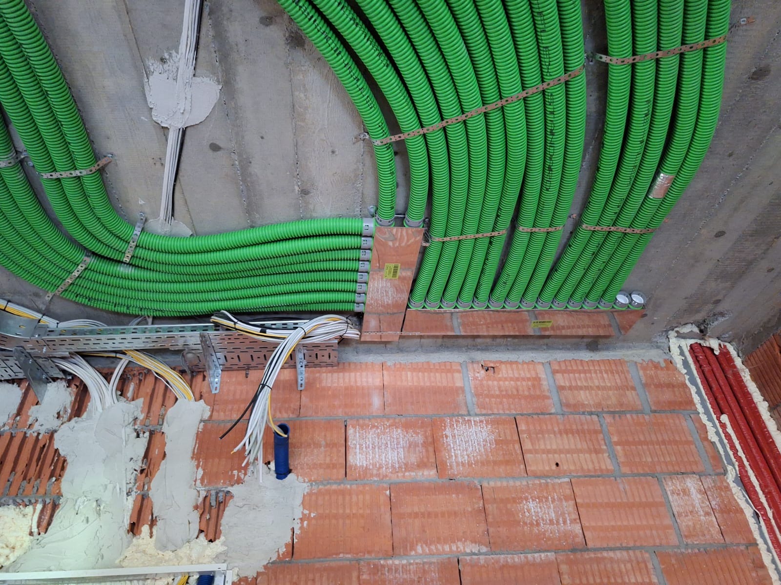 Zielone rury karbowane prowadzone pod betonowym stropem i przy murowanej ścianie z czerwonej cegły, zamocowane metalowymi obejmami, widoczne kable na metalowej drabince kablowej.