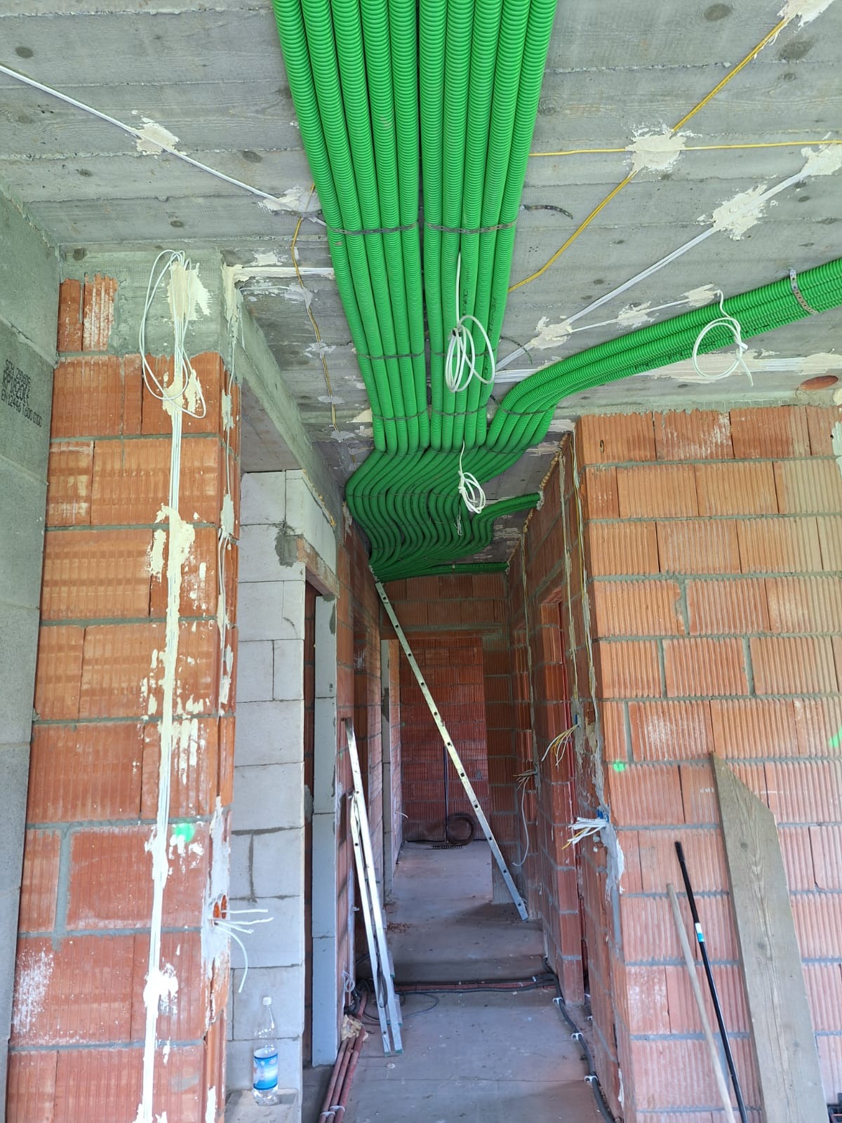 Wiązka zielonych rur karbowanych prowadzona pod sufitem w surowym stanie, z widocznymi przewodami elektrycznymi i drabiną opartą o ścianę z cegły.
