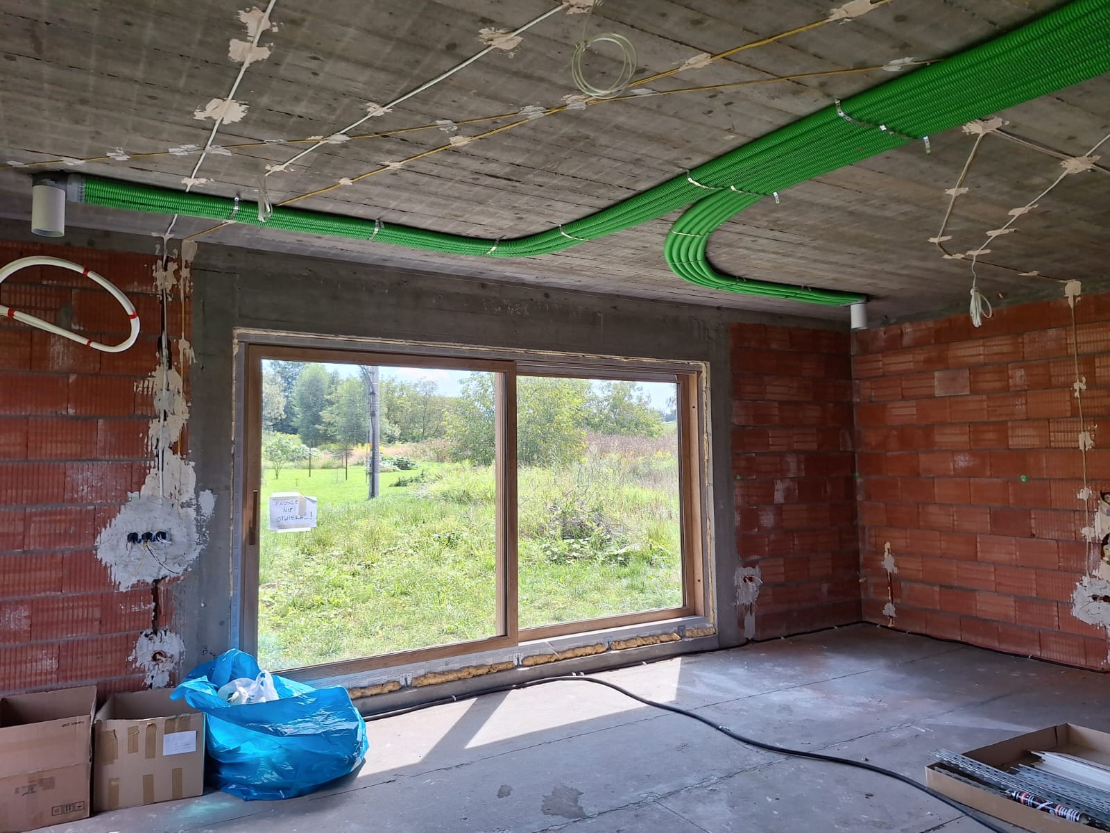 Surowy stan wnętrza z widocznymi instalacjami: zielone rury wentylacyjne pod sufitem, częściowo otynkowane ściany z czerwonej cegły, okno z widokiem na zieleń, kable elektryczne i prowizoryczne...