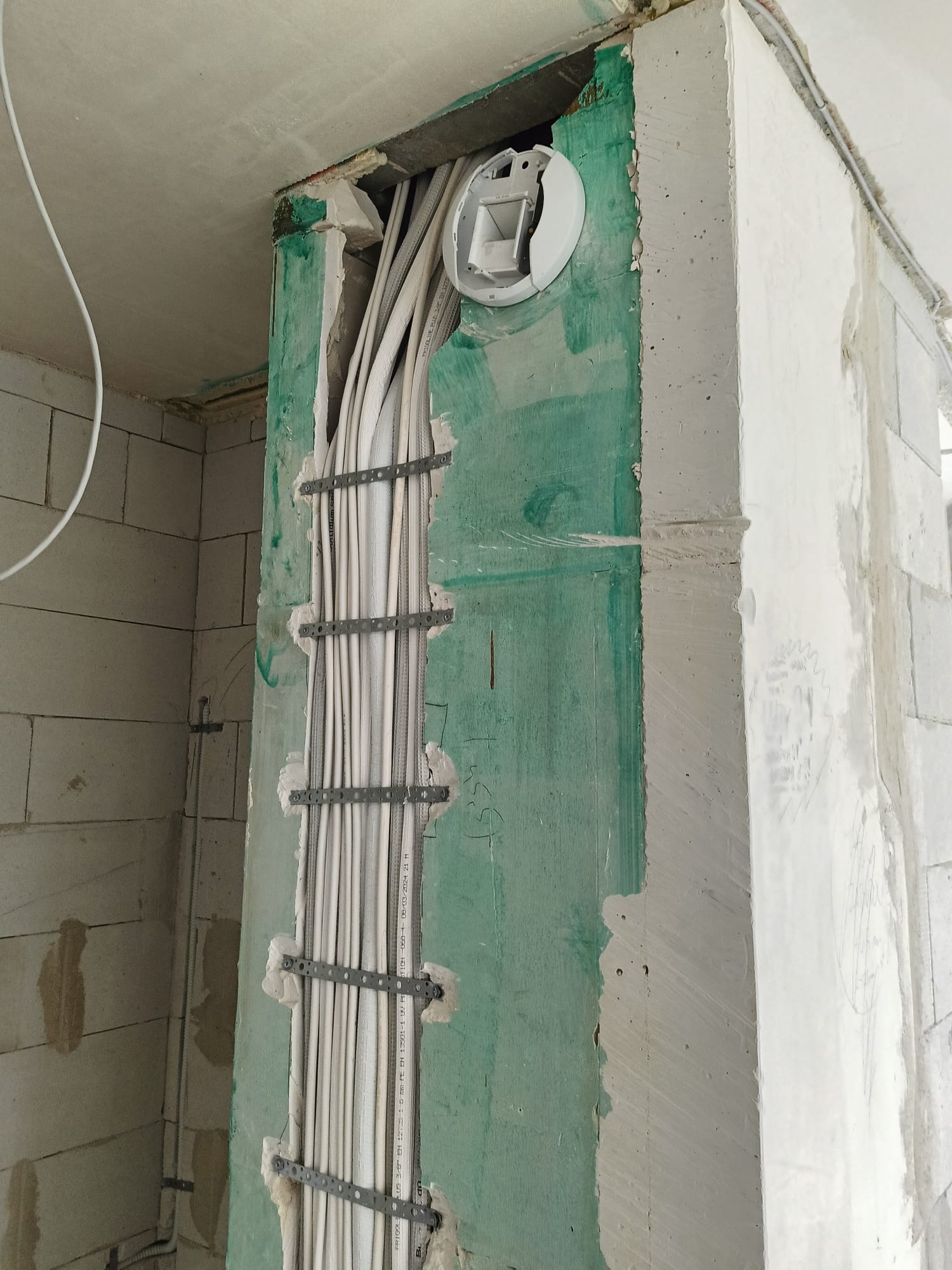 Wiązka przewodów elektrycznych zamocowana do ściany w trakcie prac instalacyjnych, widoczne wsporniki montażowe i puszka instalacyjna w ścianie.