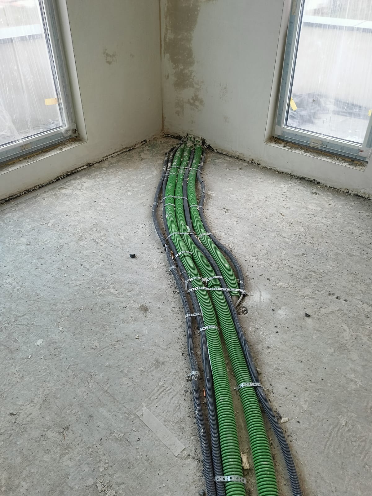 Rzędy zielonych i czarnych rur karbowanych, zabezpieczonych metalowymi obejmami, biegną po surowym, betonowym podłożu w rogu pomieszczenia z dwoma oknami.