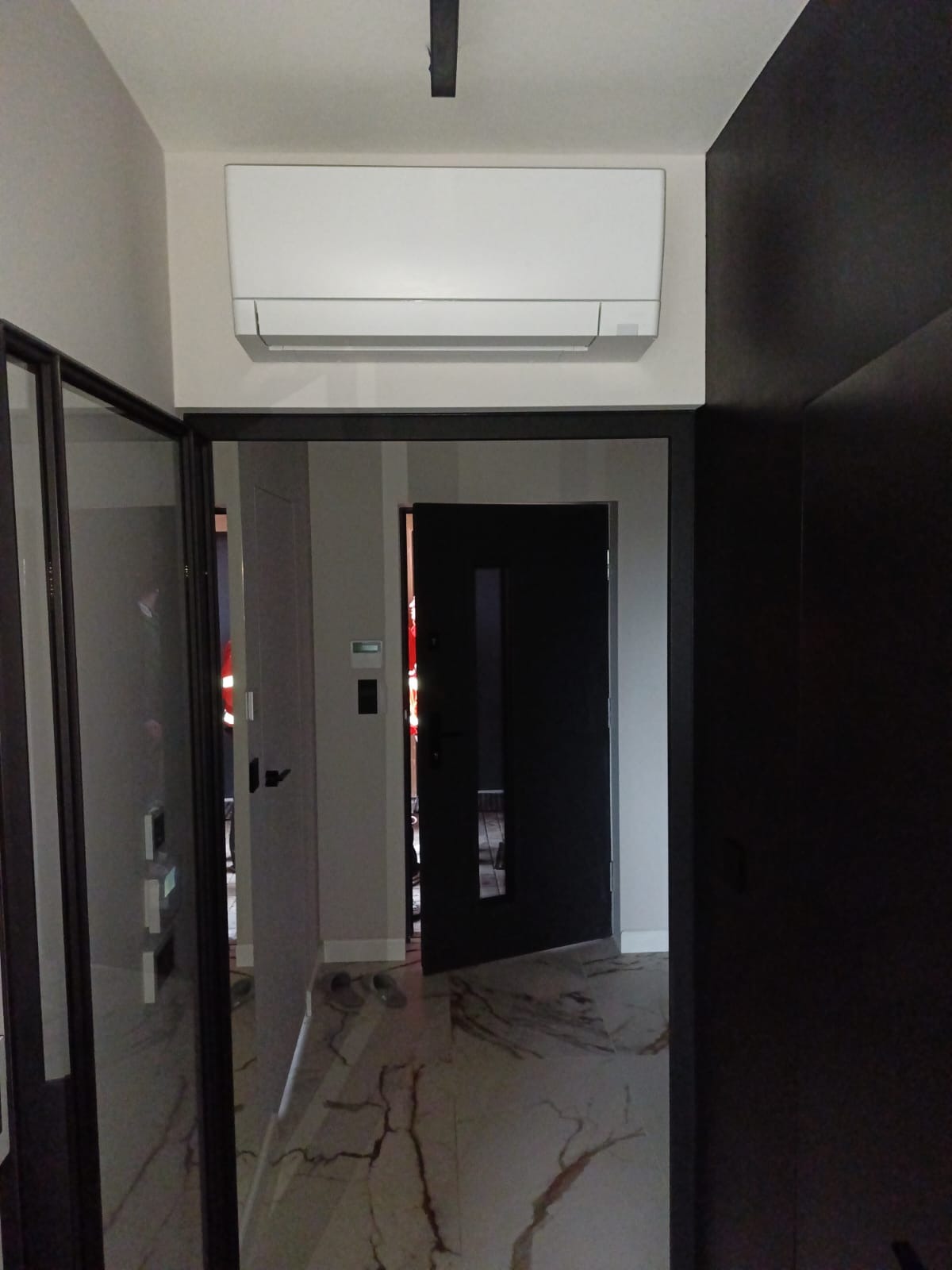 Wnętrze mieszkania z zamontowaną białą jednostką klimatyzacyjną na ścianie nad drzwiami, widok z korytarza na otwarte drzwi do kolejnych pomieszczeń, marmurowa podłoga.