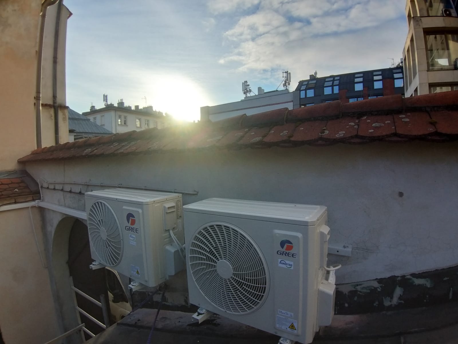 Dwa klimatyzatory marki GREE zamontowane na zewnętrznej ścianie budynku z widoczną dachówką i budynkami w tle, oświetlone wschodzącym słońcem.