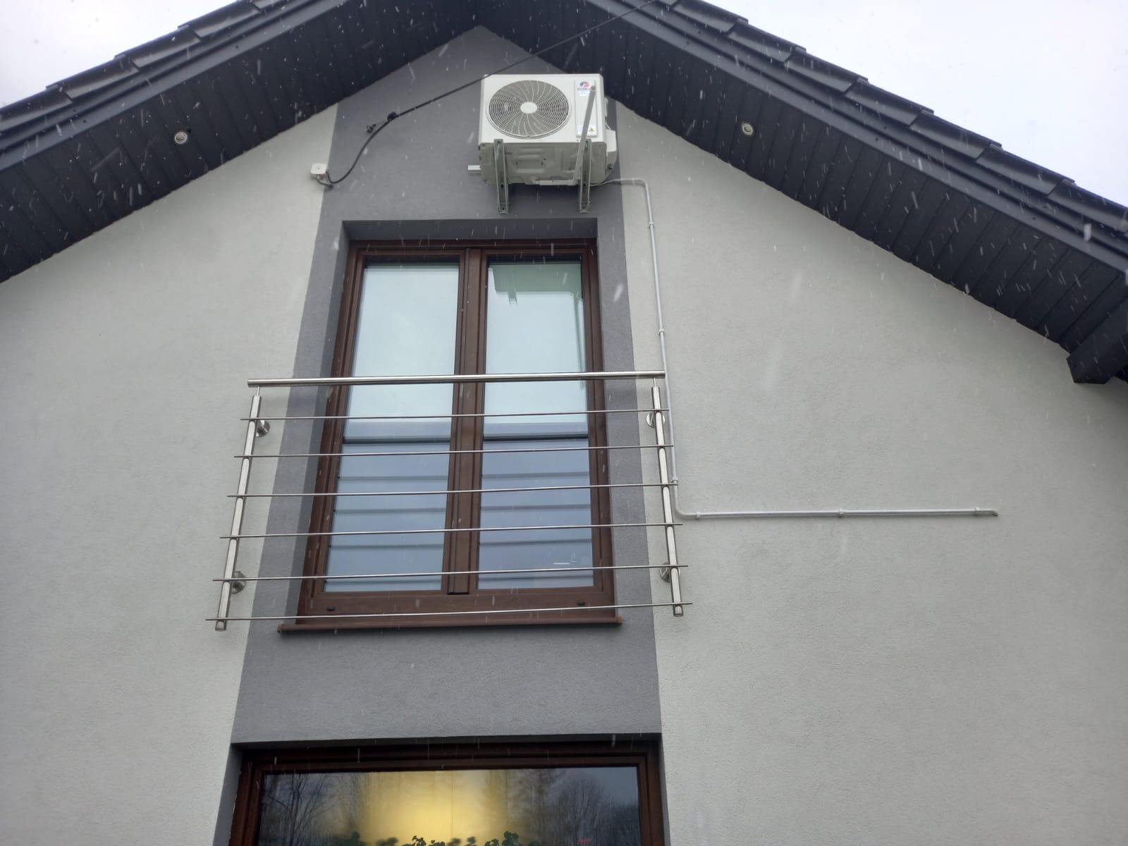 Biała jednostka zewnętrzna klimatyzatora zamontowana na elewacji domu z balkonem, widoczne rury instalacyjne i padający śnieg.