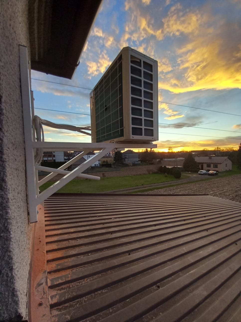 Zewnętrzna jednostka klimatyzatora zamontowana na białym wsporniku na tle zachodzącego słońca i widoku na okoliczne budynki.