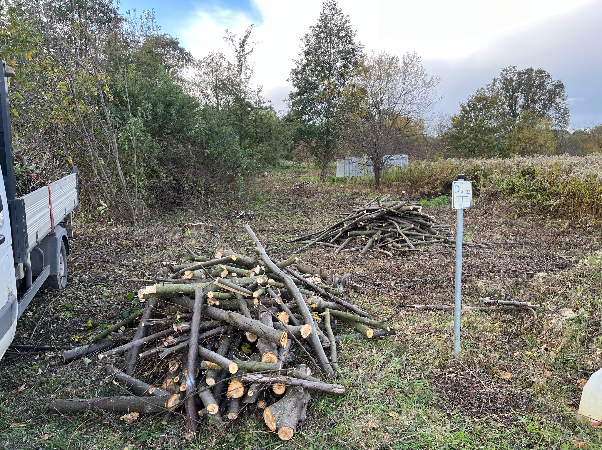 Stosy pociętych gałęzi i pni drzew leżące na trawie po wycince, widoczny biały samochód dostawczy z przyczepką oraz znak drogowy z napisem 'D o 32 T 05'.