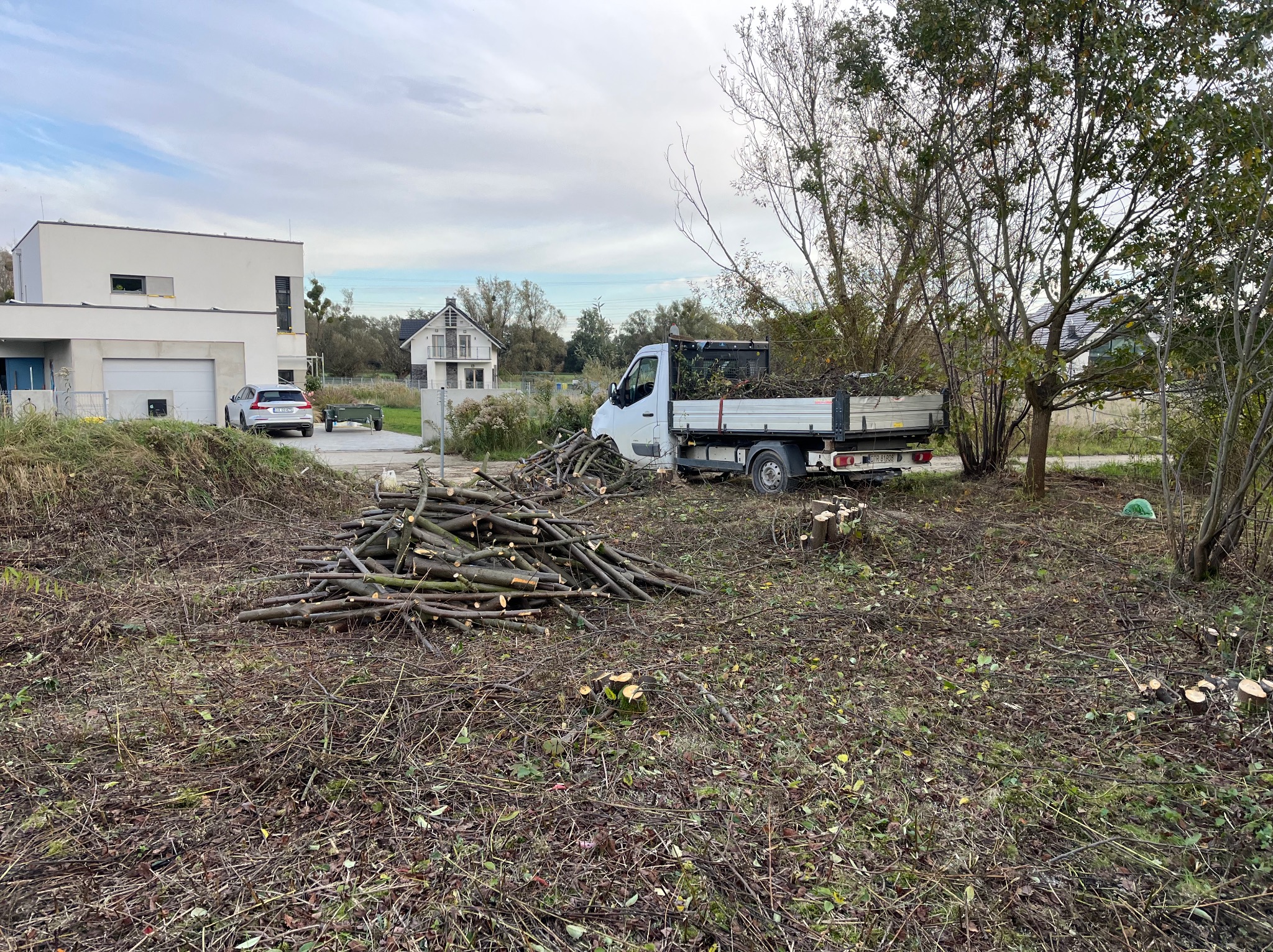 Stos pociętych gałęzi na trawie, biała ciężarówka z załadunkiem w tle, nowoczesny dom w oddali, pochmurne niebo.
