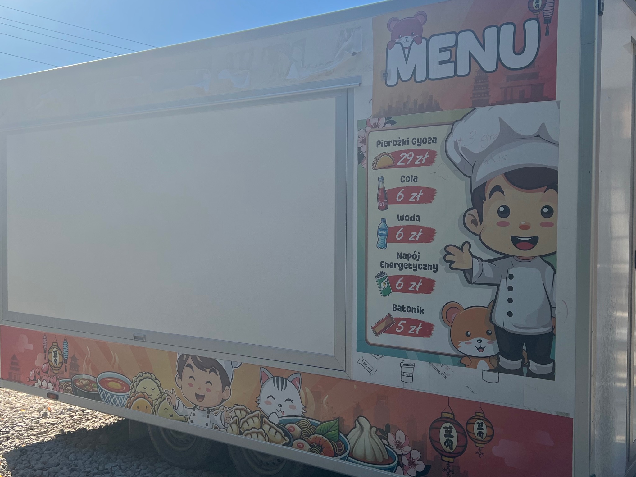 Food truck z menu pierożków gyoza, coli, wody, napoju energetycznego i batonika, ozdobiony rysunkami kucharza, zwierząt i lampionów.