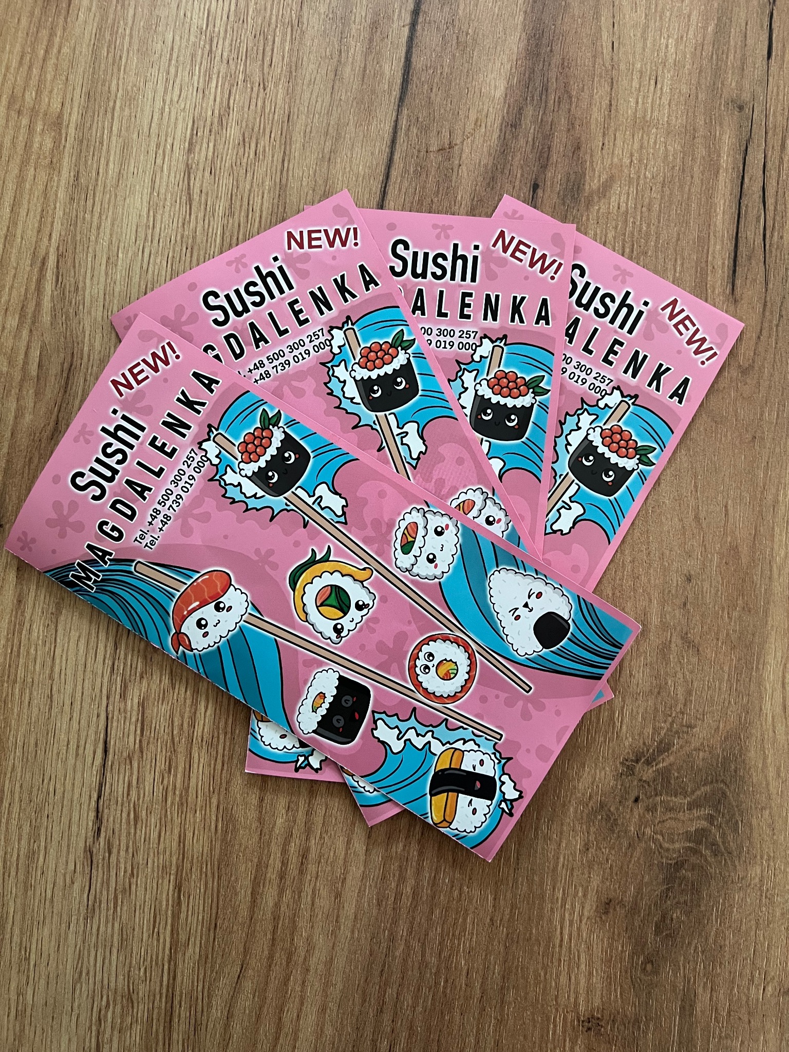 Stos różowych ulotek reklamowych sushi z rysunkowymi ilustracjami i danymi kontaktowymi na drewnianym tle.