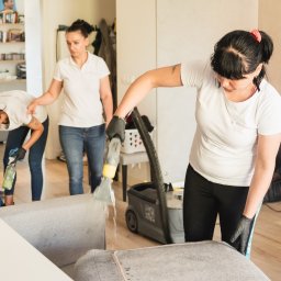 Yuliia Dovzhyk - Zespół sprzątający w Mierzynie czyści tapicerkę sofy za pomocą odkurzacza piorącego; widoczna woda wypływająca z dyszy urządzenia, a w tle koleżanka czyści podłogę i trzecia osoba nadzoruje.