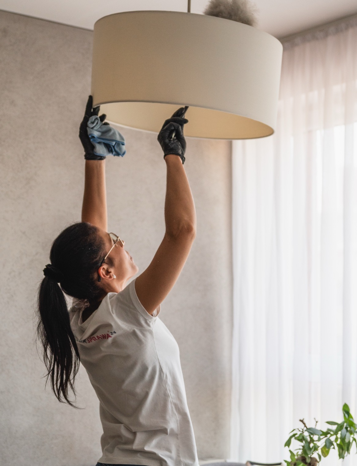 Kobieta w czarnych rękawiczkach czyści lampę sufitową szmatką, widok z dołu, jasne wnętrze.