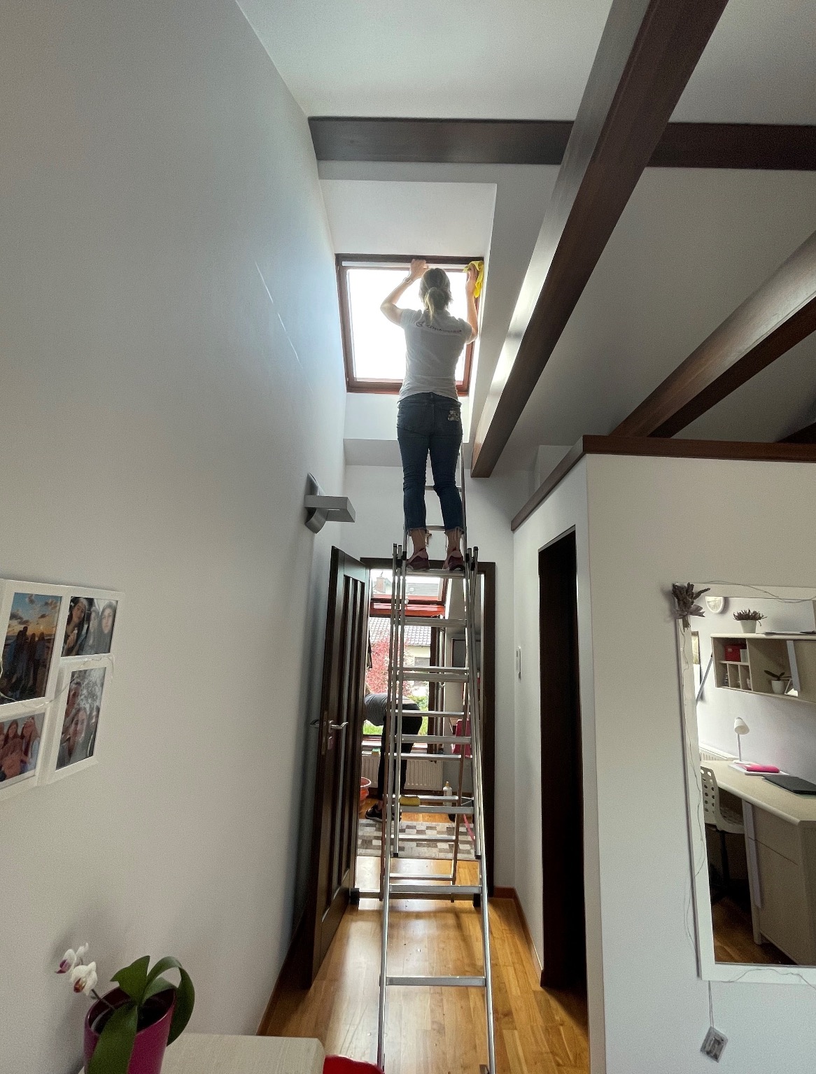 Kobieta na aluminiowej drabinie czyści okno dachowe żółtą ściereczką w jasnym wnętrzu domu z drewnianymi belkami stropowymi.