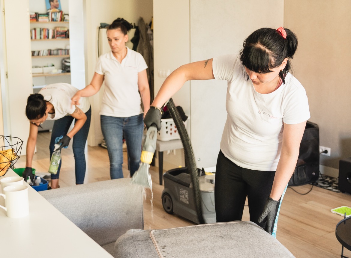 Zespół sprzątający w Mierzynie czyści tapicerkę sofy za pomocą odkurzacza piorącego; widoczna woda wypływająca z dyszy urządzenia, a w tle koleżanka czyści podłogę i trzecia osoba nadzoruje.