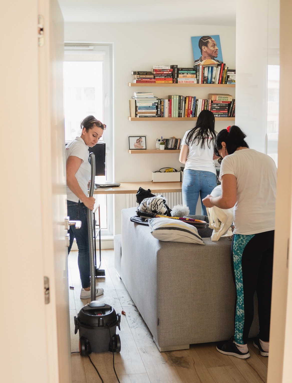 Trzy kobiety w trakcie sprzątania mieszkania: jedna odkurza podłogę, druga porządkuje rzeczy na półkach, a trzecia organizuje przedmioty na sofie. W tle regał z książkami i obraz na ścianie.