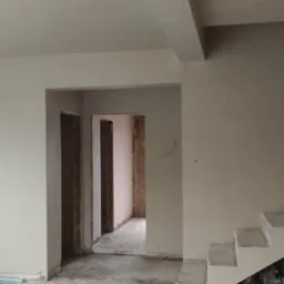 Wnętrze w trakcie prac wykończeniowych: świeżo otynkowane ściany i sufit w jasnym odcieniu, widoczne przejścia do innych pomieszczeń z surowymi ościeżnicami, częściowo przygotowane schody,...