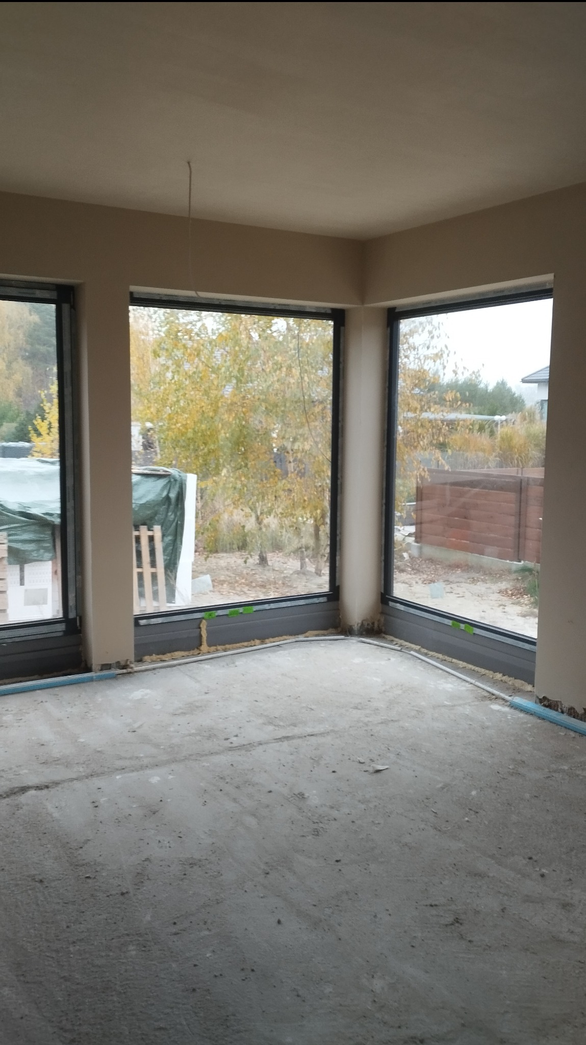 Surowe wnętrze z trzema dużymi oknami z czarnymi ramami, świeżo położone tynki na ścianach, surowa wylewka na podłodze, widoczne instalacje pod oknami, jesienny krajobraz za oknem.