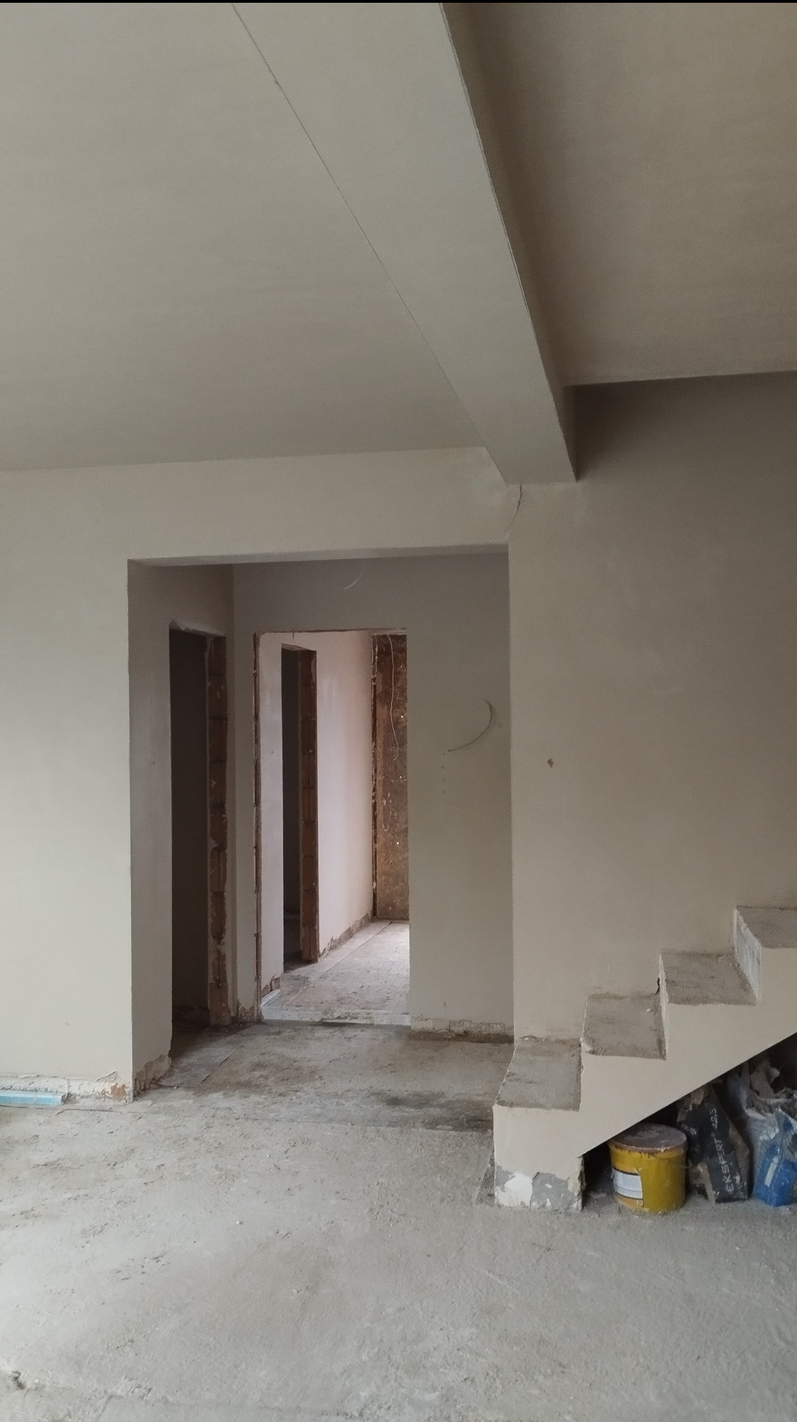 Wnętrze w trakcie prac wykończeniowych: świeżo otynkowane ściany i sufit w jasnym odcieniu, widoczne przejścia do innych pomieszczeń z surowymi ościeżnicami, częściowo przygotowane schody,...