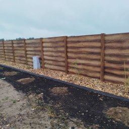 ROSMARIN Sp. z o.o. - Długi, poziomy płot z paneli imitujących drewno, z widoczną skrzynką elektryczną, oddzielający teren z przygotowanymi miejscami pod nasadzenia, widok z poziomu gruntu przy pochmurnym niebie.