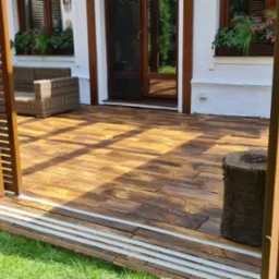 Zadaszony taras z drewnopodobną posadzką, przesuwnymi panelami z lamelami i widokiem na dom z oknami w kwiatach.