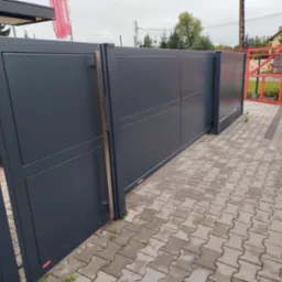 Nowoczesna, przesuwna brama w kolorze antracytowym, z widocznymi panelami i subtelnymi, pionowymi uchwytami, zamontowana na posesji z kostką brukową, z częściowo otwartym skrzydłem, ukazującym...