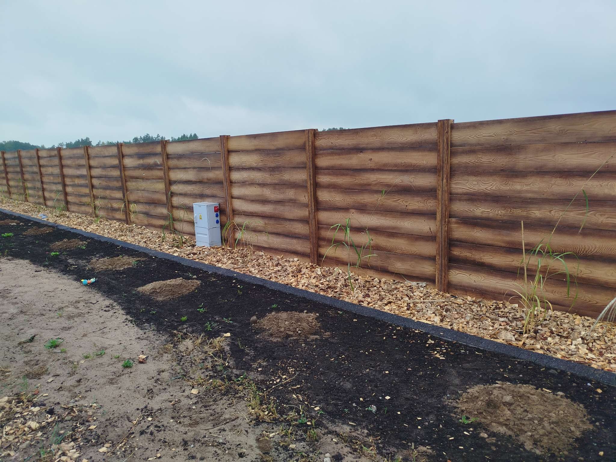 Długi, poziomy płot z paneli imitujących drewno, z widoczną skrzynką elektryczną, oddzielający teren z przygotowanymi miejscami pod nasadzenia, widok z poziomu gruntu przy pochmurnym niebie.